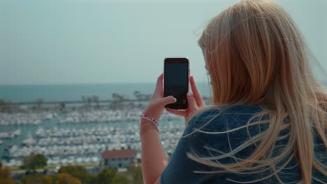 Blonde-teenage-caucasian-girl-taking-pictures-with-her-cell-phone-of-a-harbor-in-California
