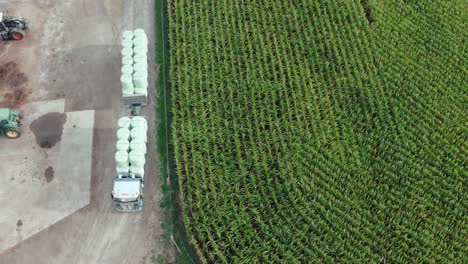 Truck-transporting-round-silage-fodder-bales-beside-corn-field-at-farm,-aerial