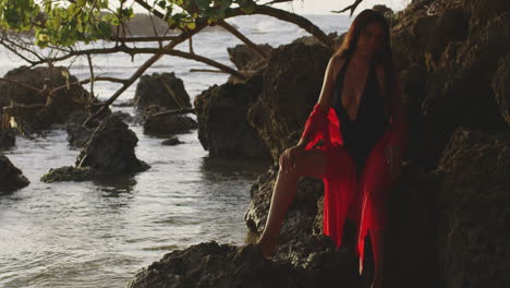 Latino-Girl-Model-in-Black,-Red-Swimwear-Poses-Sitting-on-Tropical-Beach-Rocks-at-Sunrise,-Panning-Shot