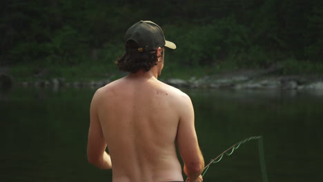 Back-of-Topless-Young-Adult-Man-Fly-Fishing,-Casting-Fishing-Line-into-Lake