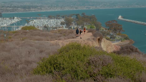 Three-people-walking-along-a-cliff-side,-dirt-trail,-above-Dana-Point-Harbor-in-California