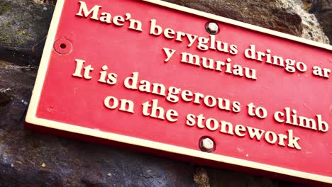 Red-metal-warning-climbing-stone-wall-sign-English-Welsh-multilingual-signage-dolly-left