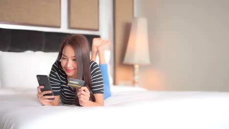 Young-attractive-Asian-woman-holding-phone-and-credit-or-debit-card-while-laying-across-bed-with-clean-white-comforter-and-pillows