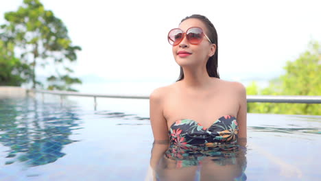 Portrait-of-an-Asian-woman-in-big-red-sunglasses-and-swimsuit-inside-the-swimming-pool-water-turning-her-sight,-slow-motion-handheld