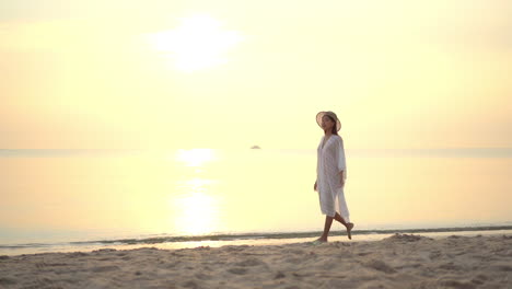 Asian-Woman-walking-on-the-beach-by-the-sea-water-on-shiny-bright-sunset-in-summer-wearing-a-white-sundress-and-straw-hat---slow-motion-static-shot