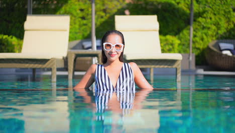 Attractive-Asian-Woman-in-Stylish-Striped-Monokini-and-Sunglasses-Relaxing-Inside-Swimming-Pool-of-luxury-Resort-Looking-Straight