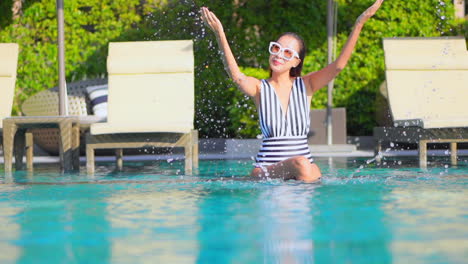 Playful-attractive-Asian-woman-playing-water-splash-smiling-sitting-at-shallow-water-of-swimming-pool-next-to-deckchairs-at-Hotel-lounge,-slow-motion