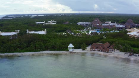 Beautiful-rising-tilting-down-aerial-drone-shot-of-the-tropical-coastline-of-playa-del-carmen-with-large-vacation-resorts-and-a-small-white-gazebo-in-Riviera-Maya,-Mexico-on-a-warm-sunny-summer-day
