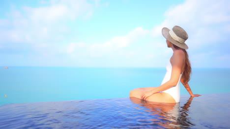 Asian-woman-in-a-white-bathing-suit-and-floppy-sun-hat-sits-on-the-edge-of-the-high-rooftop-infinity-pool-with-feet-in-the-water-together-on-a-sky-and-seascape-background