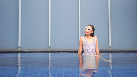 Attraktive-Asiatische-Frau-Mit-Badeanzug-Und-Sonnenbrille,-Die-Im-Urlaub-Im-Klaren,-Seichten-Wasser-Des-Resort-Swimmingpools-Sitzt