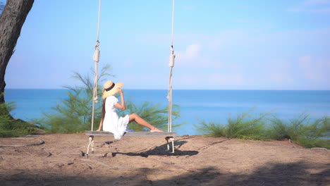 Amazing-Asian-woman-gently-sitting-on-the-big-wooden-bench-swing-wearing-summer-sundress-and-hat-on-blue-sea-background-in-Thailand,-slow-motion-copy-space