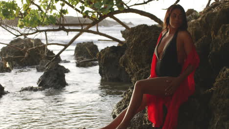 Latino-Girl-Model-Poses-Stroking-her-Legs-Skin-Sitting-on-Tropical-Island-Rocks,-Looks-at-Camera