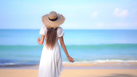 Young-Asian-Woman-traveler-in-White-Sundress-and-Straw-Sunhat-Walking-Towards-the-Sea-at-the-Picturesque-Island,-She-Raises-her-Arms-up-when-Reaching-the-Seawater,-slow-motion-back-view