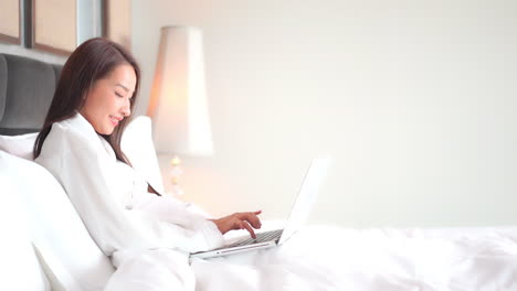 Side-view-of-Asian-woman-in-white-bathrobe-using-her-laptop-computer-lying-on-the-bed-at-the-Hotel-room,-she-is-typing-a-message-on-laptop-keyboard-smiling