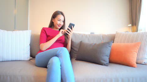 Asiatische-Frau,-Die-Ein-Handy-Benutzt,-Während-Sie-Auf-Dem-Sofa-In-Ihrem-Wohnzimmer-Liegt