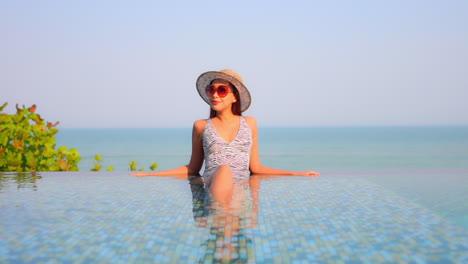 Asian-young-woman-in-sunhat-and-red-sunglasses-leaning-on-the-border-inside-infinity-pool-front-view-sunset-on-a-cloudless-sky,-template-copy-space