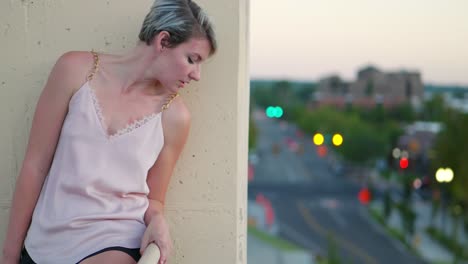 Short-haired-blonde-girl-with-dark-roots-looking-happy,-then-peeking-over-edge-of-tall-building-onto-the-city-street-at-dusk,-then-back-at-camera
