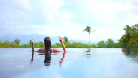 Eine-Frau-In-Einem-Swimmingpool-Mit-Dem-Rücken-Zur-Kamera-Glättet-Ihr-Nasses-Haar,-Während-Sie-Auf-Ein-Tropisches-Paradies-Blickt