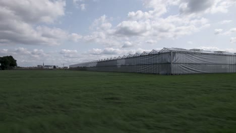 Greenhouse-in-meadow-field-seen-from-the-side-with-reflection-of-trees-visible-in-the-glass-while-approaching-sideways-and-cumulus-clouds-in-blue-sky-above