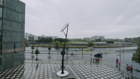 Wide-shot-of-the-front-of-Futurium-with-circle-pavement-and-river-Spree-with-moving-boat-in-Berlin,-Germany,-rainy-day