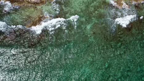 Drone-flying-over-clear-turquoise-water-arriving-to-waves-crashing-into-rocks-of-a-tropical-barren-beach-with-white-sand-and-palm-trees-in-Riviera-Maya,-Mexico-near-Cancun