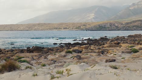 waves-crashing-on-wild-rocky-shoreline,-Elafonissi-Island-beach-on-Crete-island-with-azure-clear-water,-grassy-sand-dunes-next-to-ocean,-greece