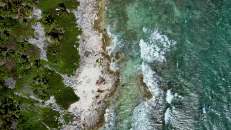 Beautiful-aerial-bird's-eye-drone-top-view-of-a-tropical-barren-beach-with-clear-turquoise-water,-white-sand,-palm-trees,-and-waves-crashing-into-rocks-near-Riviera-Maya,-Mexico-near-Cancun