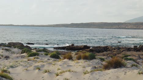 waves-crashing-on-wild-rocky-shoreline,-Elafonissi-Island-beach-on-Crete-island-with-azure-clear-water,-grassy-sand-dunes-next-to-ocean,-greece