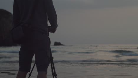 Man-Holding-Camera-On-Tripod-At-The-Beach-During-Sunset-In-Cornwall,-United-Kingdom---medium-shot