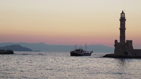old-boat-entering-the-old-venetian-harbor-with-historic-lighthouse-during-summer-evening,-sunset-ocean-view-over-chania-in-crete-greece