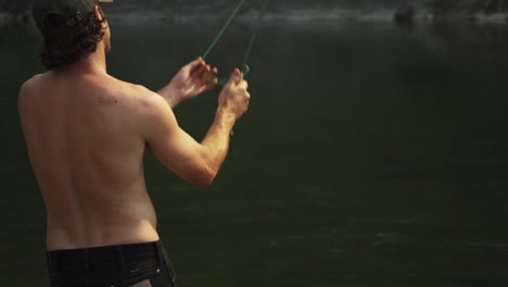 Man-without-shirt-fly-fishing-on-a-camping-trip-to-Glacier-National-Park-in-Montana,-locked-off-shot-watching-the-man-from-behind-whilst-he-is-fly-casting