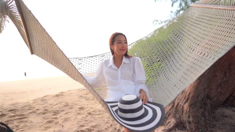 A-young-pretty-woman-rocks-gently-in-a-rope-hammock-slung-over-a-sandy-Southeast-Asian-beach
