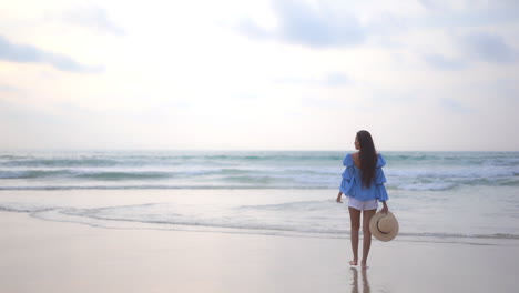 Back-to-the-camera,-slow-motion,-rule-of-thirds,-an-attractive-woman-in-shorts-and-a-bohemian-blouse-holds-a-sunhat-in-one-hand-as-she-walks-into-the-oncoming-surf
