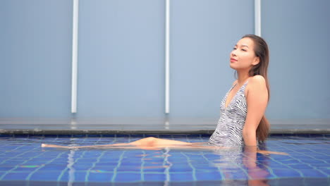 Thailändische-Frau-In-Zebrafarbenem-Monokini,-Die-Im-Flachen-Schwimmbadwasser-Sitzt-Und-Die-Beine-Unter-Der-Wasserzeitlupe-Bewegt,-Urlaubsvorlage