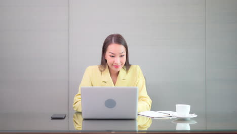 Stylish-Asian-businesswoman-sitting-at-the-work-desk-in-front-of-her-laptop-computer-and-typing-on-the-keyboard-with-a-happy-face-expression,-front-view-slow-motion