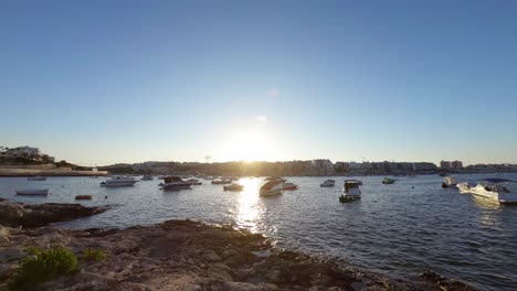 Malta,-Qawra,-Salina-Bay-Bei-Sonnenuntergang-Mit-Schwimmenden-Booten-Und-Beleuchteter-Stadt-Im-Hintergrund
