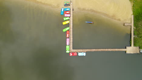 Aerial-Panning-Top-down-View-People-Walking-Along-the-Wooden-Jetty-with-Pedal-Boats-Moored-Near-the-Pier-in-Hartowiec-Lake,-Poland