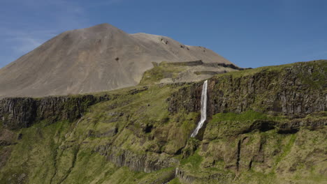 Pintoresca-Cascada-De-Islandia-En-Un-Acantilado-Con-Una-Montaña-Volcánica-En-El-Fondo