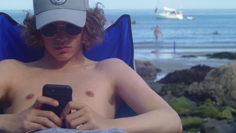 Curly-haired-Man-Sitting-And-Using-Smartphone-At-Coverack-Beach-In-Cornwall,-UK