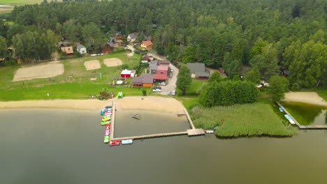 Aerial-Orbiting-Around-Hartowiec-Lake-Waterfront-Resorts-and-Rental-Buildings-for-Summer-Vacation,-Volleyball-Field-and-Shore-in-Green-Landscape,-Wooden-Piers-with-Docked-Boats-on-Forest-Background