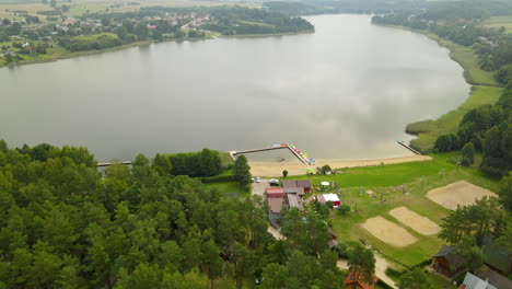 Aerial-tilt-down-from-Hartowiec-lake-landscape-to-Private-Hotels-equipped-for-summer-water-sports-like-kayaking,-canoeing-pedal-boarding,-and-fishing-surrounded-by-green-dense-forest,-Poland