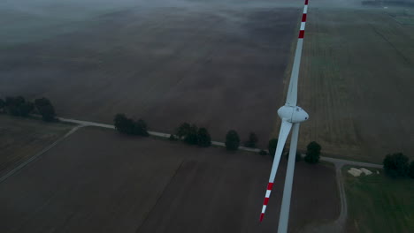 Aerial-close-up-of-slow-rotating-wind-turbine-on-brown-farm-field-in-nature---Production-of-renewable-energy-during-windless-early-morning-in-dawn