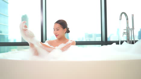 Asian-Young-Woman-Playing-with-Foam-in-the-Bathtub-at-High-floor-Apartment-with-Glass-Wall-and-City-view,-Girl-is-Spreading-Foam-on-her-Body-and-Arms