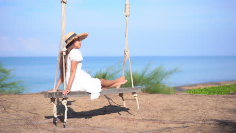 Young-Woman-in-Straw-Sunhat-and-White-Dress-Sitting-and-Swinging-on-a-Wooden-Bench-Swing-Hanging-on-a-Tree-at-a-Tropical-Beach-in-Thailand-Enjoying-the-Sea-View,-slow-motion