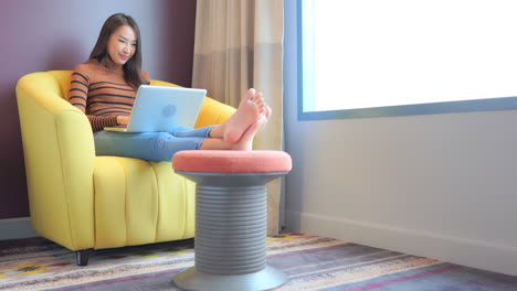 Asian-Woman-Typing-on-Laptop-Keyboard-While-Sitting-in-Yellow-Armchair-Having-her-Legs-Resting-on-the-Puff-at-Apartment-Room