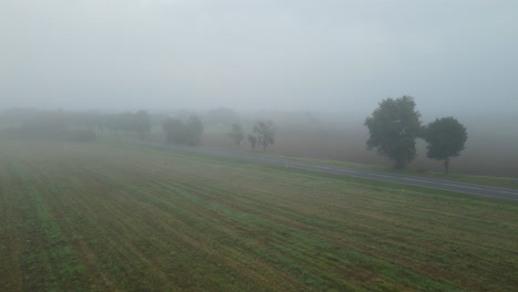 Aerial-view-over-farm-field-in-fog-showing-driving-car-on-rural-road-in-autumn