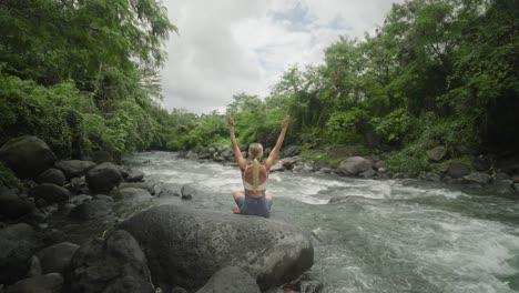 Behind-blond-fit-woman-raising-arms-while-in-easy-pose-on-rock-beside-tropical-river