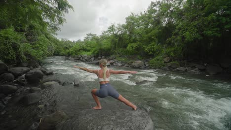 Woman-practicing-warrior-pose-on-rock-along-calming-flow-of-water-in-jungle