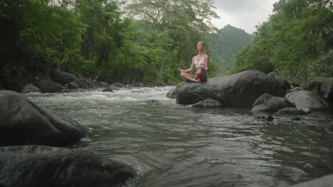 Woman-in-yoga-Sukhasana-lotus-pose-on-rock-next-to-river,-soul-searching