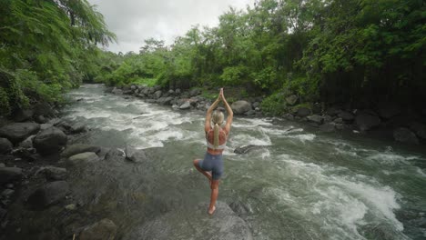 Cuerpo-Tonificado-Femenino-Practicando-La-Pose-Del-árbol-Vrikshasana-En-Una-Roca-Al-Lado-Del-Río,-Bali